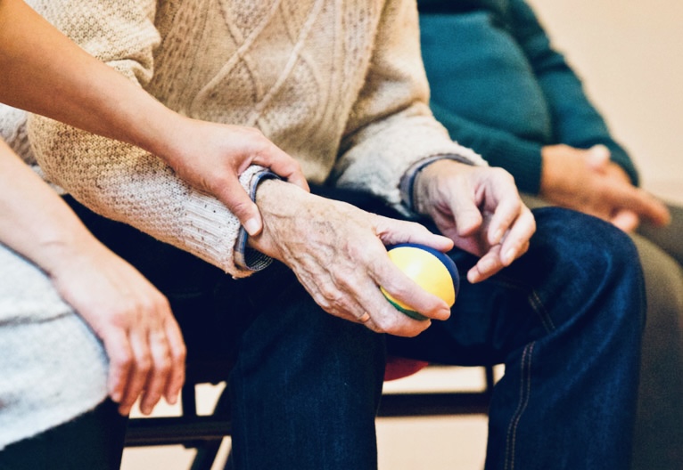 Eine Hand legt sich auf das Handgelenk eines älteren Menschen. Der Ältere hält einen kleinen Ball.